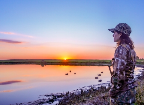 A woman waterfowl hunting by a wetland at sunset