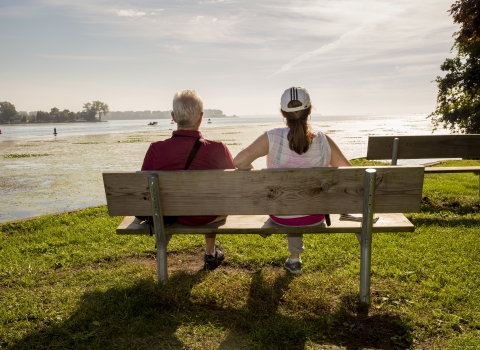 A man and a woman sit on a bench, with their backs to us, facing the river at Detroit River International Wildlife Refuge.