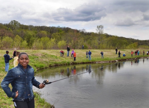 A young person with a fishing rod looks at us from edge of a body of water, with others along shore in the distance 