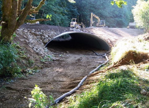 Dry streambed with new culvert for fish passage installed, with heavy machinery on the road above