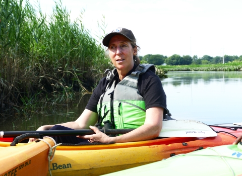 woman in lifevest in kayak in water near a shore with tall grasses