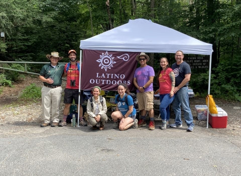 a group of people stand by an outdoor tent. A flag raised on the tent says "latino Outdoors" with a symbol of a sun above it. THe people are smiling wearing hats, binoculars and backpacks