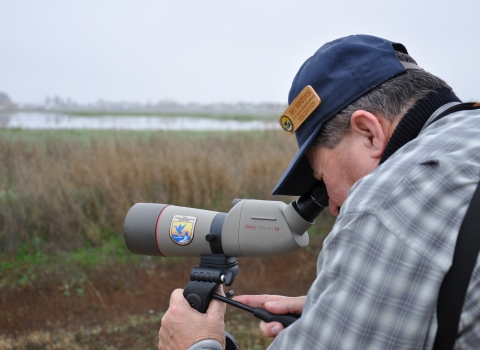 A man in a cap peers through a scope aimed at tall grass around a lake
