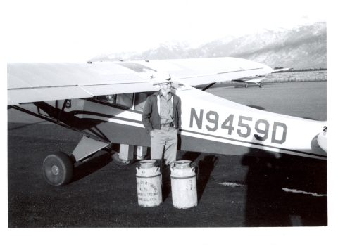 A man stands next to a small aircraft with milk cans of fish to release