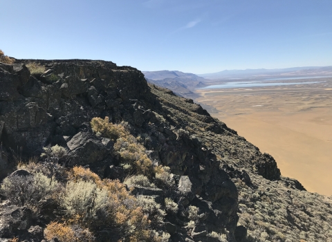 Rocky cliff faces covered in sagebrush rise above a valley floor full of fields and wetlands.