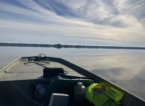 The prow of a boat looks out over a glassy lake surface reflecting cloudy sky and forests on the horizon.