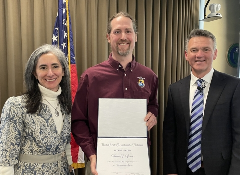 Three people smile standing in front of an American flag, and the man in the middle holds an award