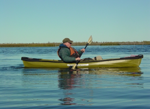 A man in an olive-green colored kayak on the water with blue sky, trees and brush in the distance.