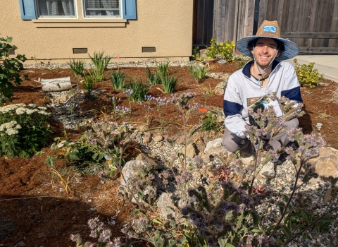 a man wearing a wide brimmed hat squats among plants in a front yard.