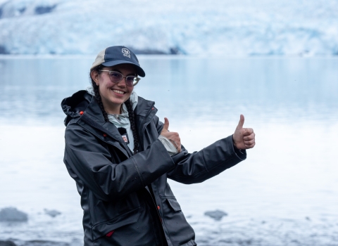 A young woman in a dark rain jacket smiles and holds up two thumbs up. She stands in front of icy water, with a large glacier visible in the background.