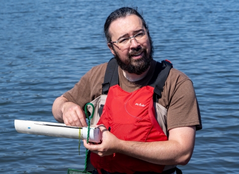 A service intern wearing waders and a PFD smiles as he measures the length of a fish. Water can be seen in the background. 