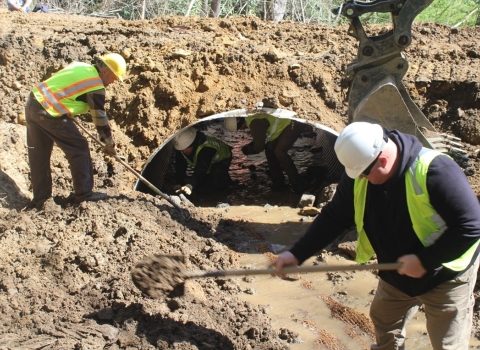 Two people in hard hats and vests shovel dirt in front of a metal culvert.