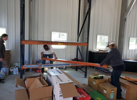 Three people building industrial shelving. Background is evenly spaced windows at chest height. Foreground has a variety of partially opened boxes of varying size