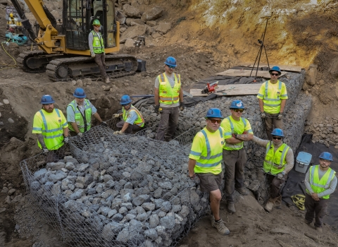 Ten people wearing bring yellow shirts and blue hard hats pose near a newly constructed rock wall