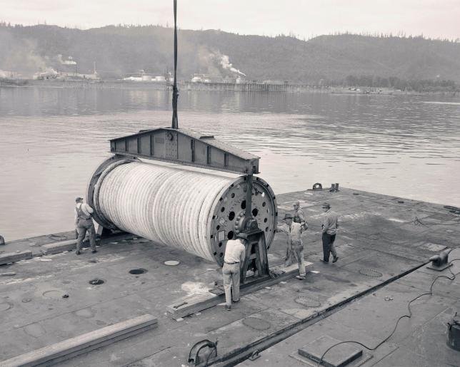 Black and white photo of large spool on dock with several workers nearby.