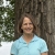 a white woman with blond hair and blue eyes, wearing glasses and a light blue polo shirt, in front of a tree, with a lake in the background