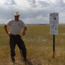 A man in a cowboy hat stands next to a signpost and fence in a grassy field.