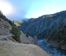 A river runs alongside a road. Trees and mountains in the distance. 