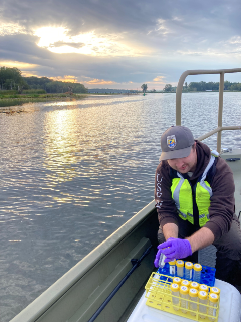 USFWS staff on a boat picks up a tube from a rack for eDNA sampling under a cloudy sky.