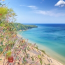 View from a sandy bluff overlooking a turquoise-blue lake, with clear water fading to deep blue toward the horizon. Green forested shoreline curves into the distance under a bright blue sky with a few wispy clouds, while leafy shrubs and branches frame the foreground.
