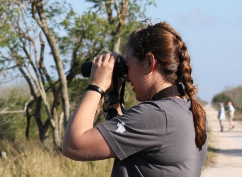 A woman with a braid in her hair looking through binoculars at a scrubby landscape