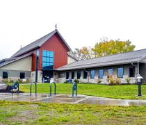 Photo is taken from the ground looking up on a new facility with stone and red siding under a blue and white sky with green grass in foreground
