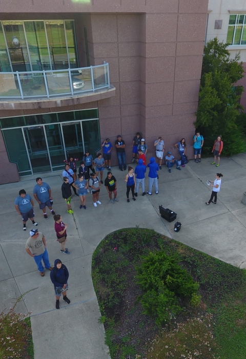 An aerial photograph of a group of people standing on the sidewalk outside of a large building