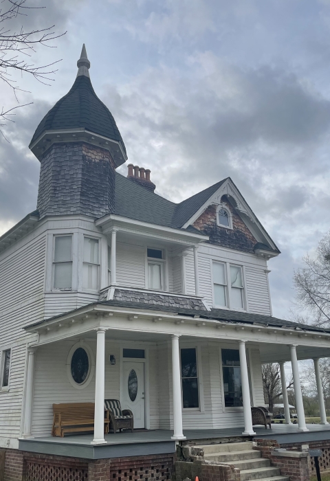 White two-story house with a front porch, nine columns, and a dark roof in the foreground with grey skies behind it.