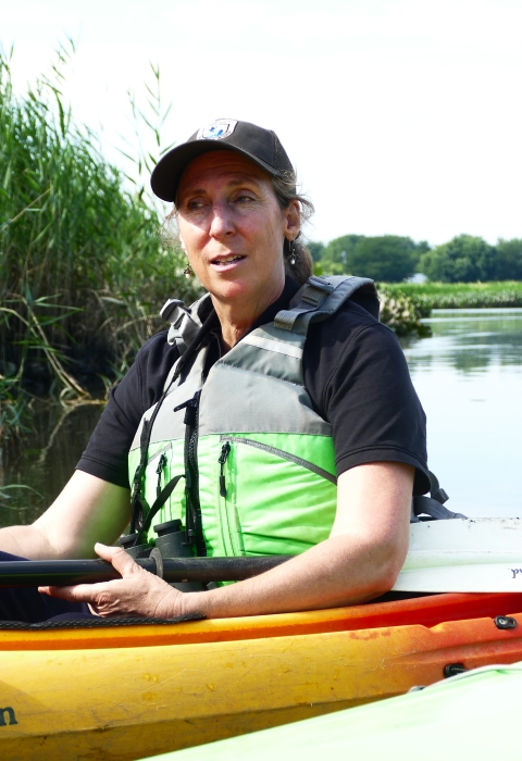 woman in lifevest in kayak in water near a shore with tall grasses