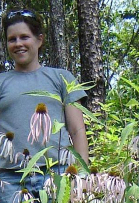 Heather Alley stands and smiles amongst tall smooth coneflower plants. The plant's petals are purple. 