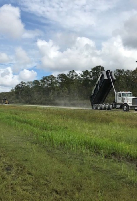 A dump truck tips a load of gravel onto a gravel road bordered in field and forest.