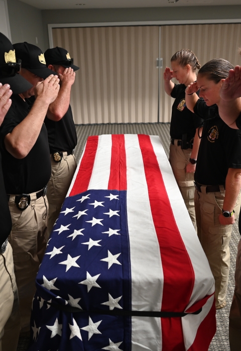 The honor guard stands over a coffin draped in an American flag. Each of them is saluting and looking down at the coffin/flag.