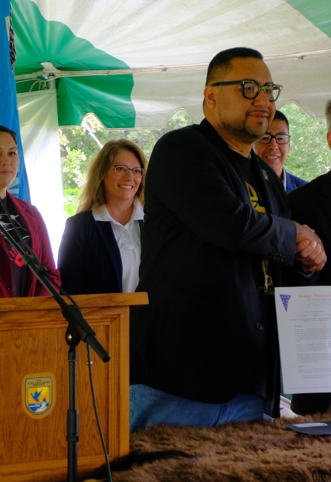 Five people stand behind two men who are shaking hands and hold up documents