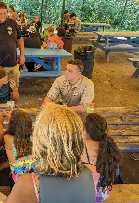a group of kids and a Service employee sit and talk at a picnic bench