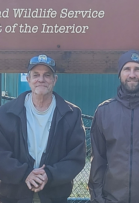 Four men stand smiling in front of a large sign for the Eagle Creek National Fish Hatchery. Sunlight and green grass are behind them.