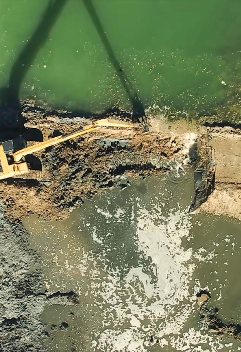 A bulldozer works to remove part of an earthen levy separating two bodies of water, while a group of people watch. 