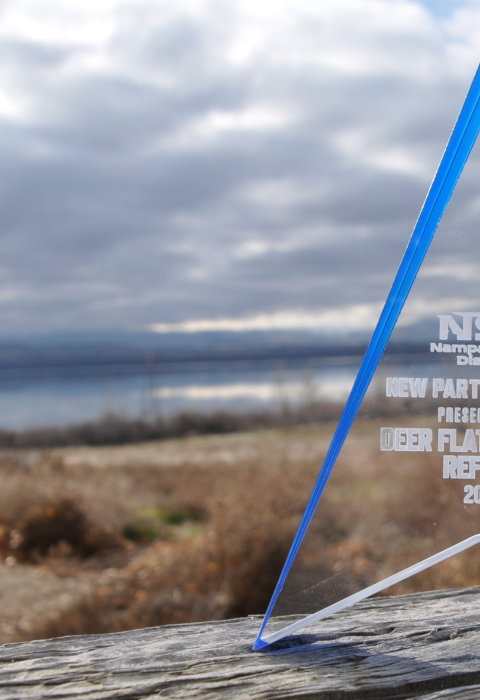 A translucent rectangular award sits in front of a lake and sagebrush habitat with an engraving that reads “Nampa School District New Partner Award Presented To Deer Flat National Wildlife Refuge 2023”