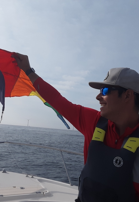 A person wearing a red shirt, black flotation vest and tan cap holds a yellow, red and orange kite aloft while standing in the bow of a boat