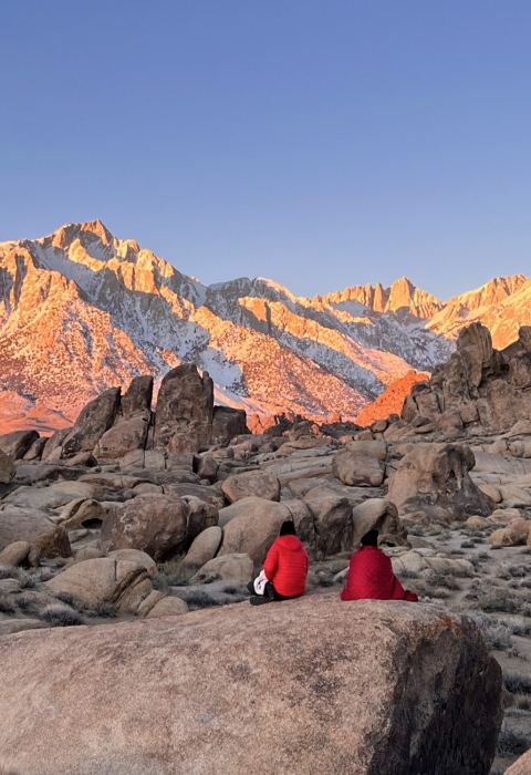 Two people sit on a large rock in the foreground of the picture, with their backs facing the camera as they stare off toward sunrise on the Eastern Sierrva Nevada.