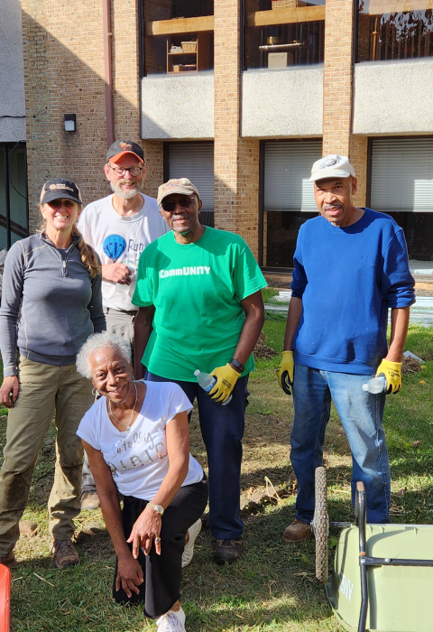 Five people stand in the yard of a building with wheelbarrows and other gardening tools