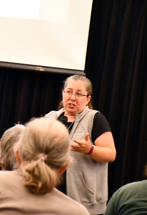A woman speaks in front of a group of older people in front of a photo screen and a black curtain.