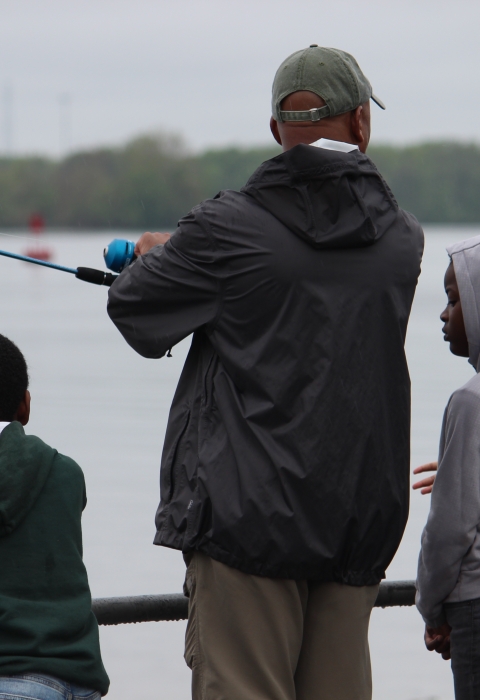 A man stands on the waterfront with a fishing rod surrounded by kids