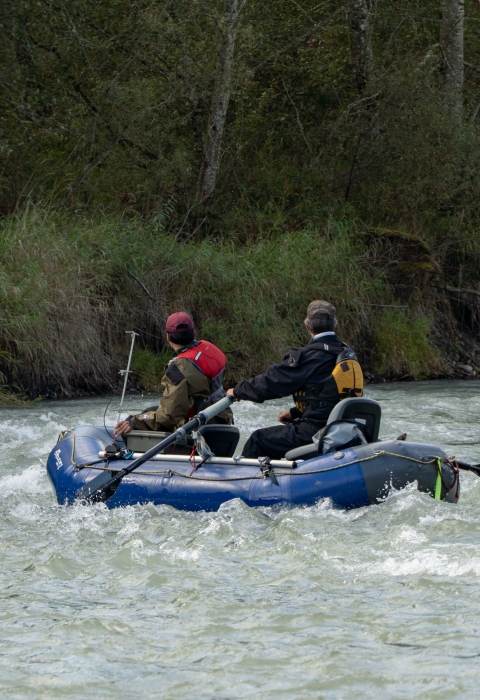 A service intern and USFWS employee float down a river in a two-man inflatable raft. They are wearing dry suits and PFDs, trees and ground foliage can be seen in the background.