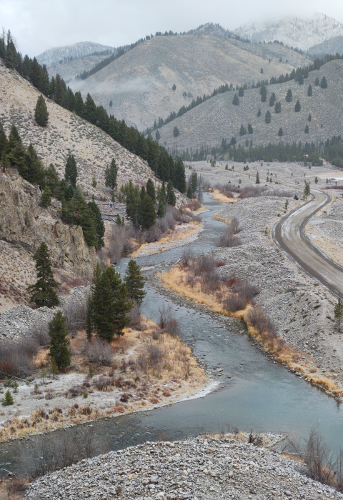 An aerial view over a winding river with a road along one side and mountains with evergreen trees on the other. there is a light dusting of snow on the road and terrain.