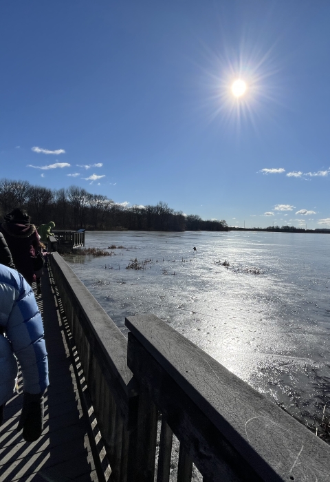 Sun shining over people walking on a boardwalk suspended over a frozen body of water.