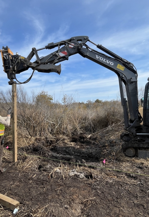 three people stand near a pole in the dirt as a small excavator pounds it into the ground