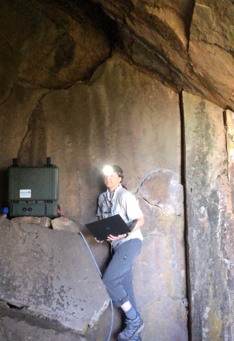 a woman with a headlamp stands in the entrance of a cave holding a laptop