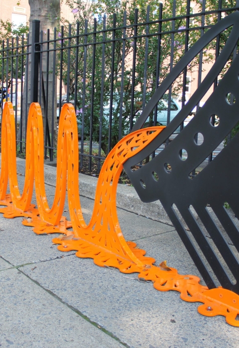 a metal art installation of orange waves pouring from a black can sits on a sidewalk, with a black metal fence and green shrubs in the background