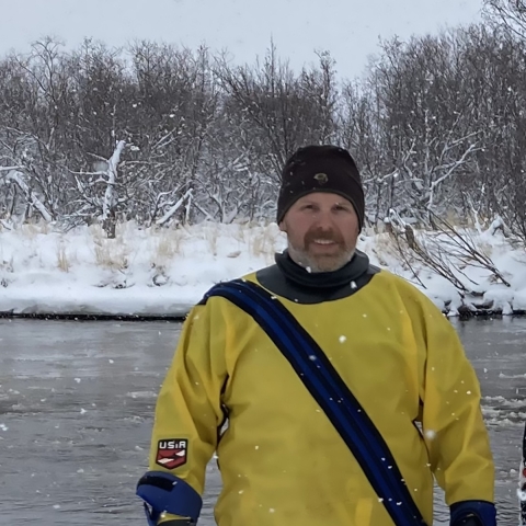 A person wears a dry suit and smiles as they stand on the snowy shore of a river.