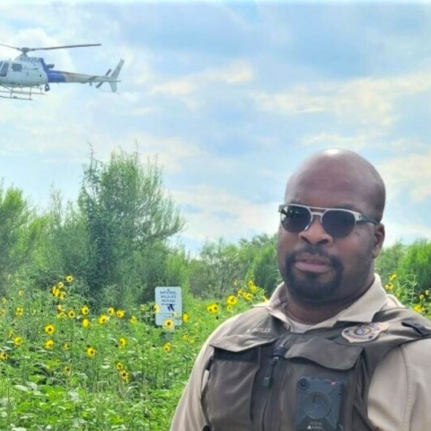 A man in sunglasses wearing a U.S. Fish and Wildlife Service uniform standing in a green field with a helicopter overhead